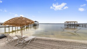 Una playa con sillas de descanso y una sombrilla de paja, pasarelas de madera sobre el agua, y cielos despejados.