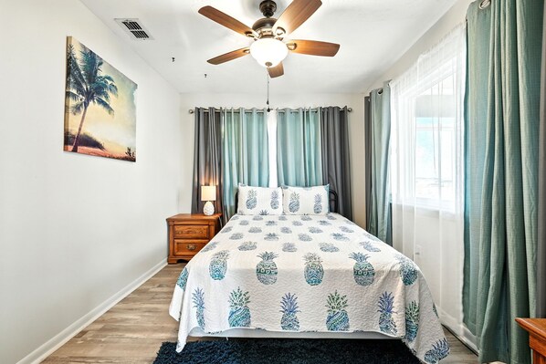Un dormitorio con cama, ventilador de techo, mesita de noche, lámpara y un cuadro de una palmera.