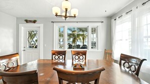 Un comedor con mesa y sillas de madera, una araña y ventanas amplias con vistas a palmeras y una piscina.