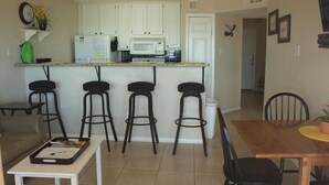 A compact kitchen with white cabinets, a microwave, and a refrigerator. There are four black bar stools and a wooden dining table with chairs.