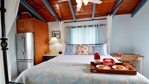 Ein Schlafzimmer mit einem Bett, Holzboden, Deckenventilator, Nachttisch, Lampe, Kühlschrank, Korbsessel und einem Frühstücksbrett auf dem Bett.