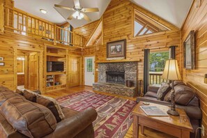 A log cabin interior with a stone fireplace, leather sofas, a television, and a ceiling fan.