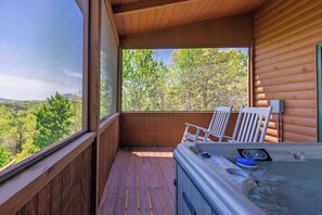A wooden deck with a hot tub, two white chairs, and a view of a forested area.