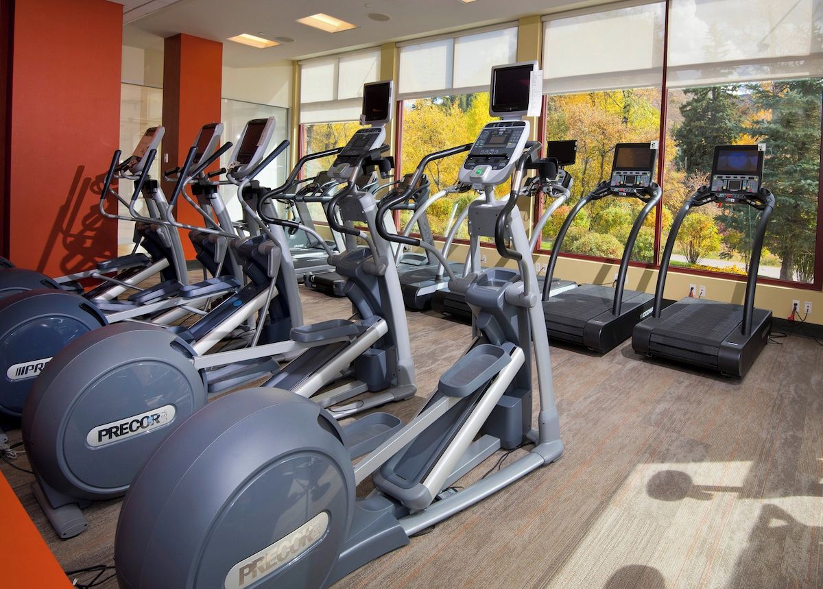 Ein Fitnessstudio mit mehreren Laufbändern und Ergometer, großen Fenstern mit Blick auf Bäume und einem Teppichboden.