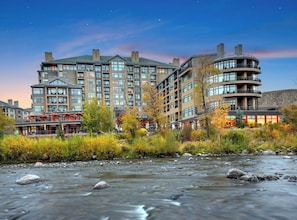 A multi-story hotel with a river in the foreground, featuring a modern architectural design with balconies and large windows.