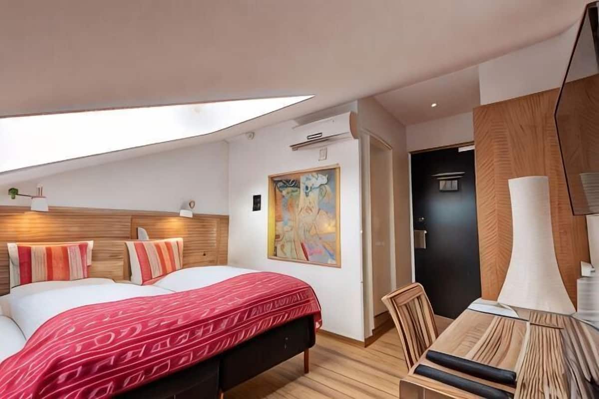 Une chambre d’hôtel moderne avec une lucarne, un lit avec une literie à motifs, un bureau avec une lampe et un grand tableau au mur.