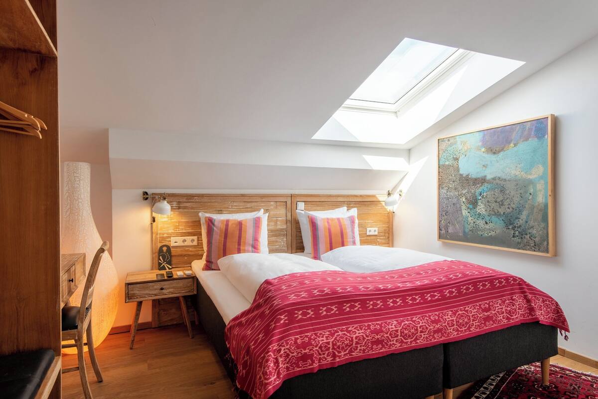 Une chambre avec une lucarne, un lit recouvert d’une couverture à motifs, une table de chevet avec une lampe et un tableau accroché au mur.