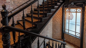 Una escalera con barandillas negras ornamentadas y escalones de madera que conduce a una habitación con un amplio ventanal y una lámpara colgante.