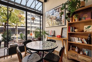 Un espace intérieur accueillant avec une table ronde et des chaises, une bibliothèque remplie de livres et un panneau, ainsi qu’une grande fenêtre donnant sur la rue.