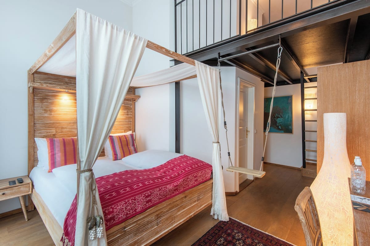 Un lit à baldaquin avec une couvre-lit rouge à motifs, une tête de lit en bois et une chaise suspendue en forme de balançoire.