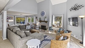 Un salón amplio con sofá gris, sillones de mimbre, chimenea y un reloj grande en la pared.