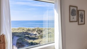 Una habitación con un amplio ventanal con vistas a una playa, enmarcada por cortinas blancas.