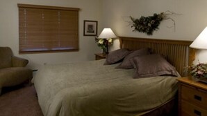 Ein Schlafzimmer mit einem Bett, einem Nachttisch mit Lampe, einem Sessel und einem Fenster mit Jalousien.