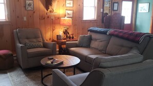 Ein gemütliches Wohnzimmer mit Holzwänden, einem aufgehängten Hirschgeweih, einem Sofa, einem Sessel, einem Couchtisch und einer Lampe.