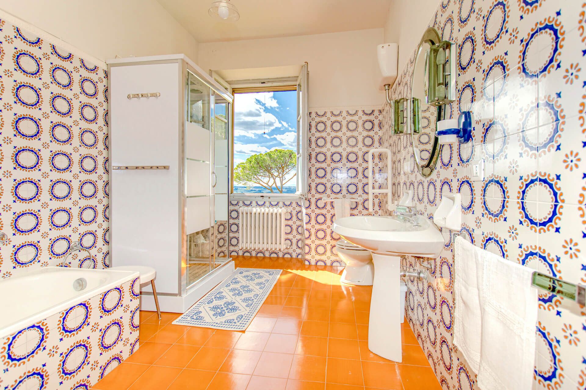 Une salle de bain avec des carreaux décorés, une baignoire, un lavabo et une fenêtre donnant sur des arbres.