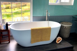 Un baño moderno con una bañera independiente, una cesta de mimbre y vistas a un paisaje verde a través de una ventana.