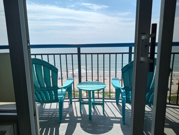 A balcony with turquoise chairs and a small table, overlooking a beach with people and umbrellas. - "Direct" Ocean Front 9th Floor cozy coastal cottage Reasonable Rates (Myrtle Beach)