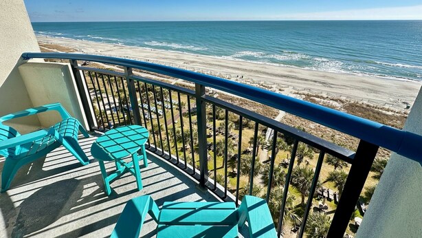 A balcony with turquoise chairs and a table overlooking a beach with a clear view of the ocean. - "Direct" Ocean Front 9th Floor cozy coastal cottage Reasonable Rates (Myrtle Beach)