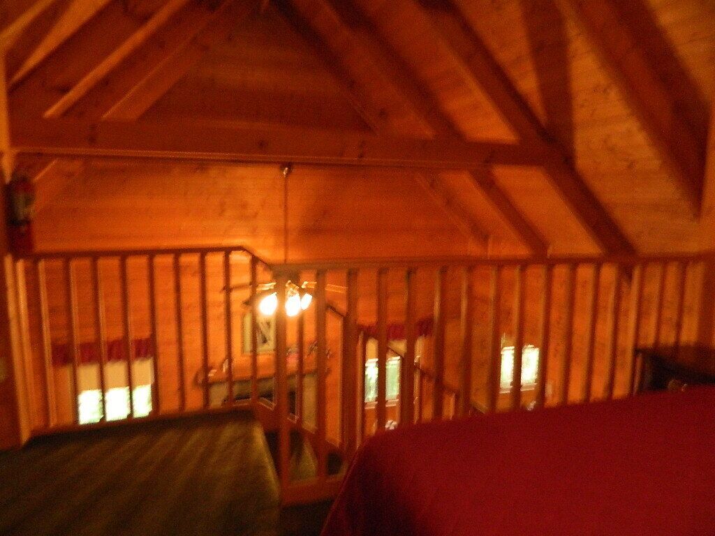 A wooden cabin interior with a sloped ceiling, a bed, and a small table with a chair.
