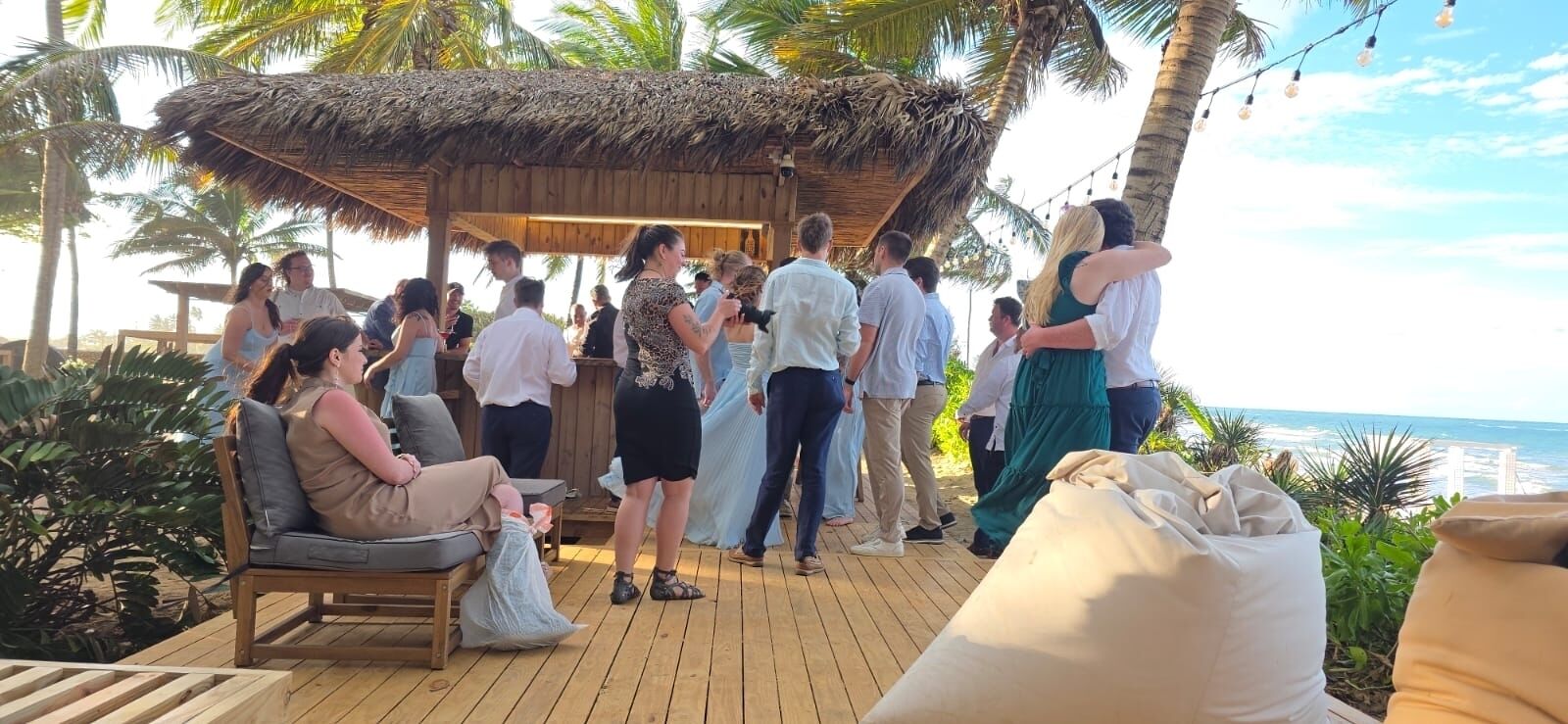 A wooden deck with a thatched-roof structure, surrounded by palm trees and a view of the ocean.