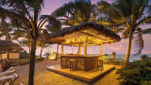 Un bar situé en bord de mer, doté d’un toit de chaume, d’une terrasse en bois et de tabourets de bar.
