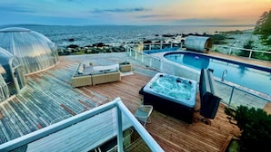 A deck with a hot tub, lounge chairs, and a glass-enclosed area overlooking the sea. - Relaxing Oceanfront Retreat . Hot tub -sauna-pool-clear domes-private (South Shore)