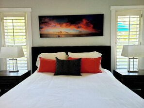 Een slaapkamer met een groot bed, twee nachtkastjes, een schilderij boven het bed en ramen met jaloezieën.