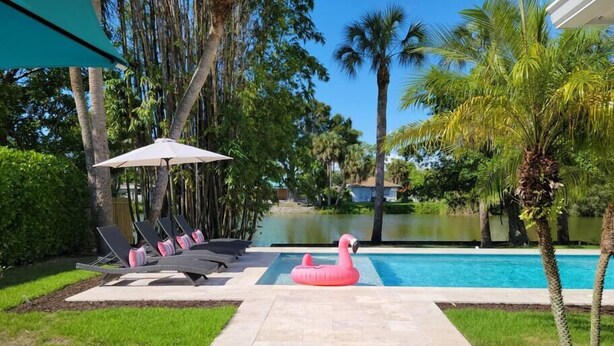 A pool area with lounge chairs, a pink flamingo float, and palm trees. - 841 Vanderbilt Beach Bliss • Lux Waterfront Home (Naples)