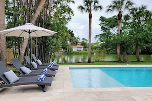 A poolside area with lounge chairs and an umbrella, surrounded by palm trees and a view of a body of water.