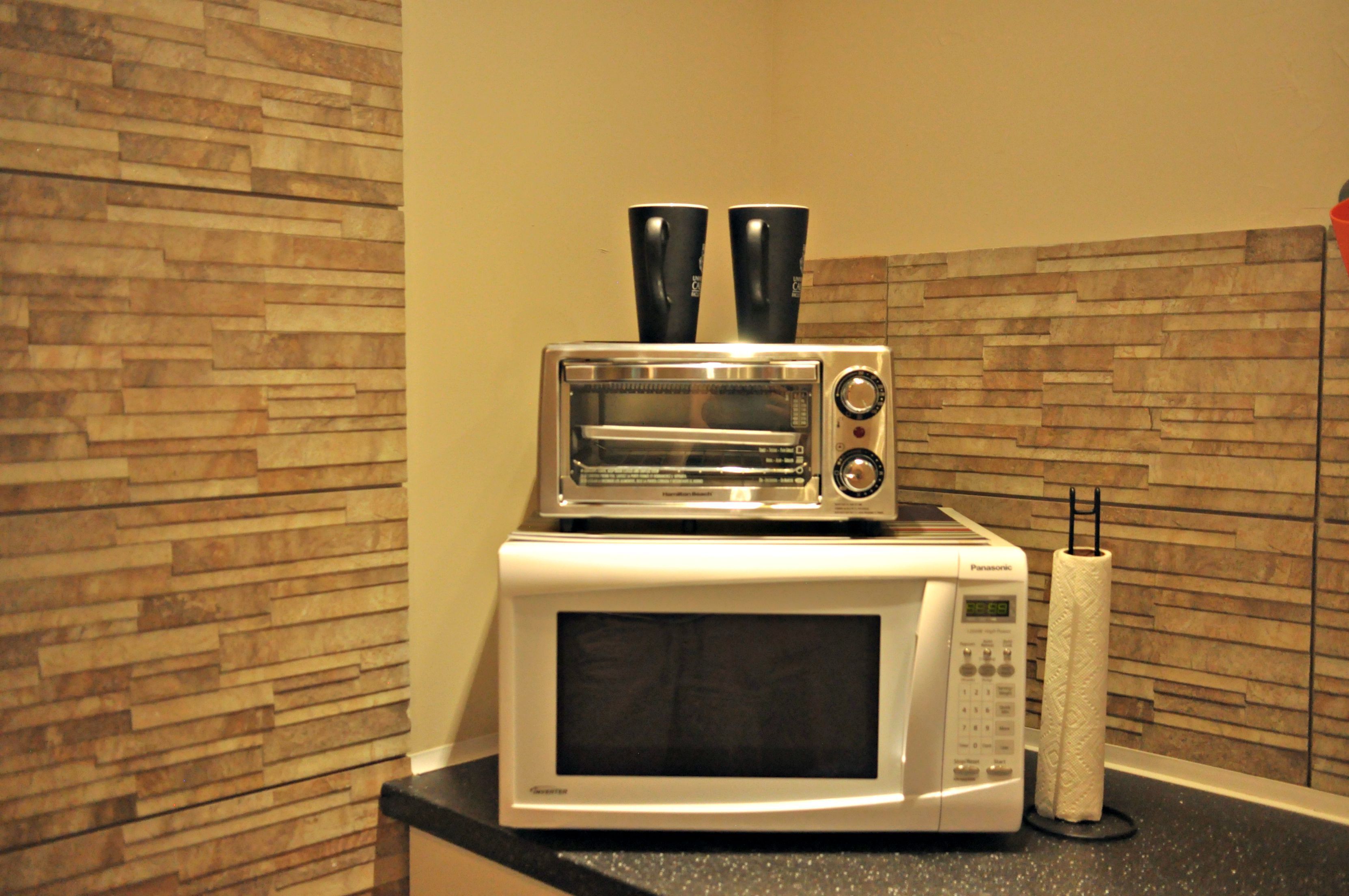 A kitchen with a microwave and a toaster oven stacked on top of each other, a paper towel holder, and a stone-patterned backsplash.