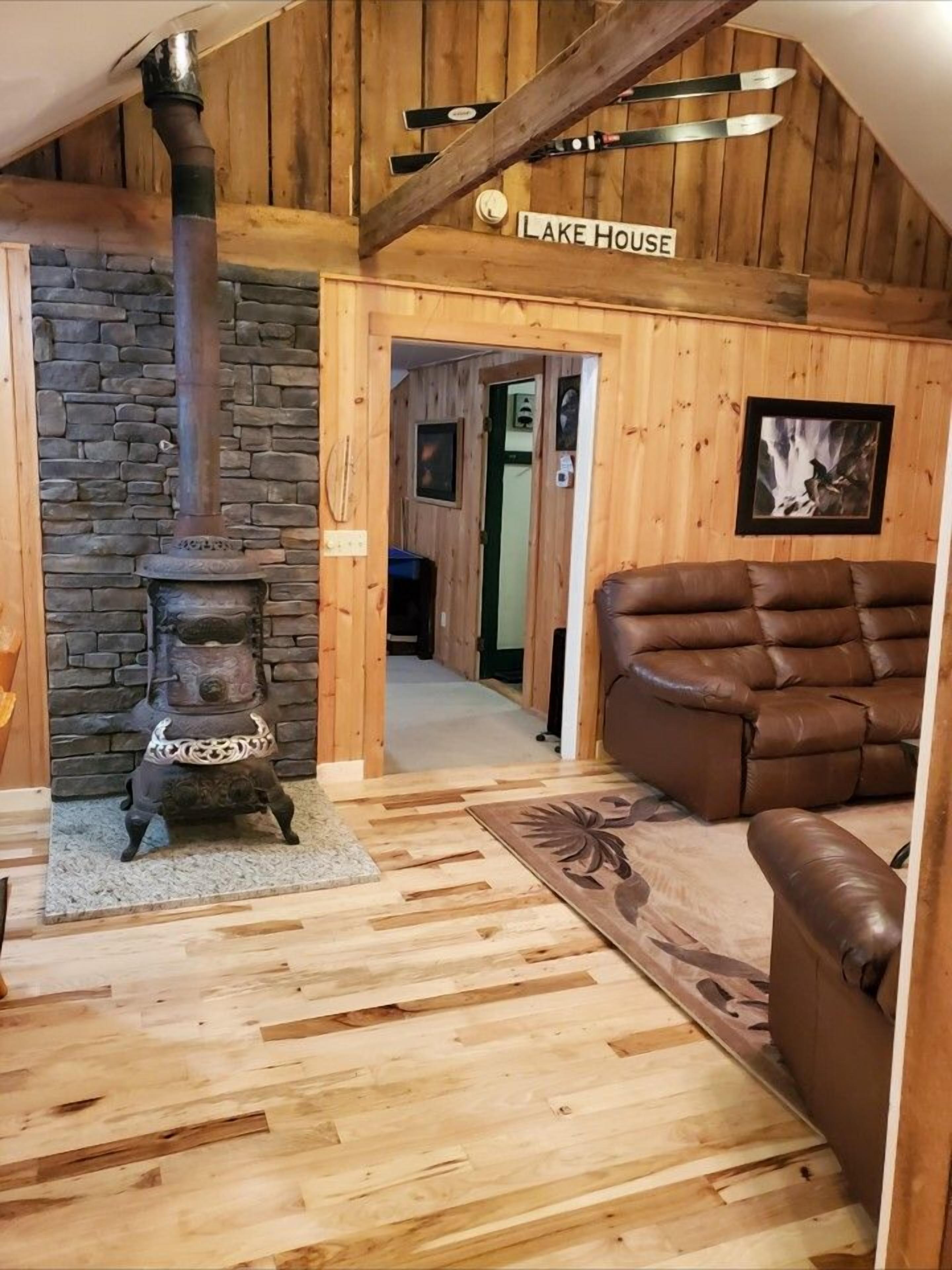 Un interno rustico con camino in pietra, pavimento in legno, un divano in pelle e un cartello con la scritta 'Lake House'.