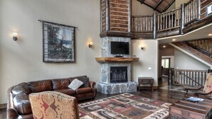 A spacious living room with a leather sofa, patterned rug, and a stone fireplace. - Castle Glen Dream Estate! Spa, movie theater, lake and mountain views! (Big Bear)