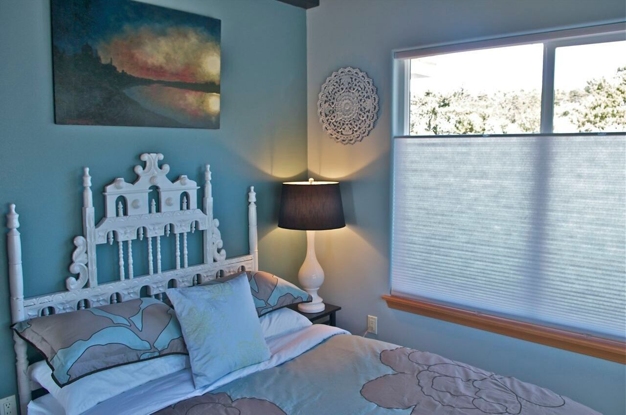 A bedroom with a white ornate headboard, a bed with patterned bedding, a bedside lamp, a decorative wall piece, and a window with blinds.
