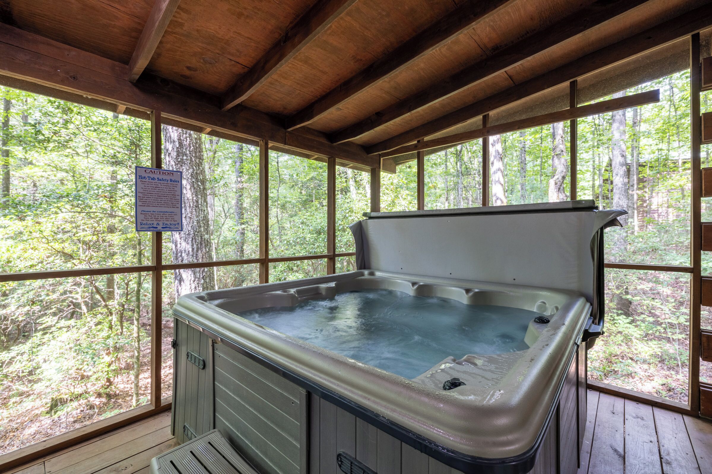 A hot tub in a wooden cabin with large windows, a caution sign, and a view of the forest.