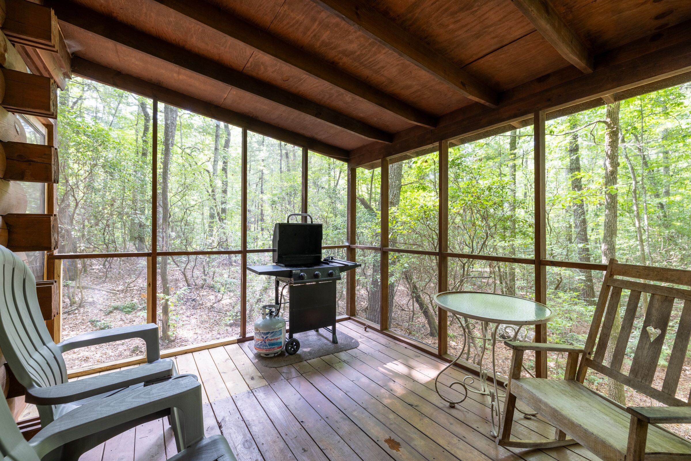 A covered porch with wooden flooring, a grill, a table, and two Adirondack chairs.