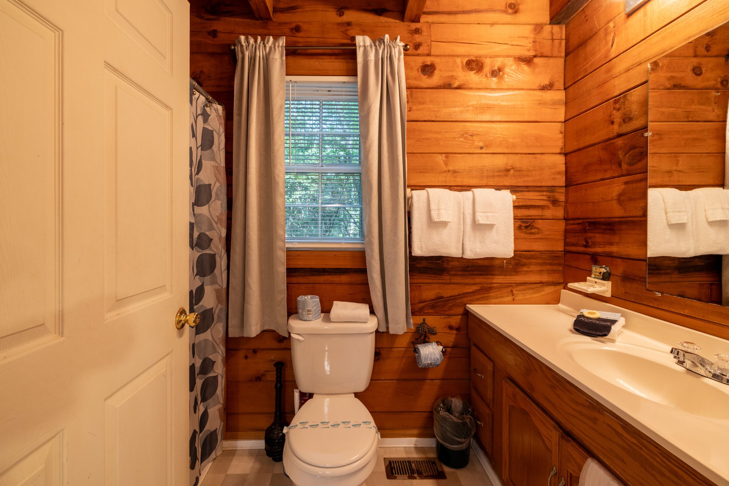 A wooden cabin bathroom with a toilet, sink, and window with curtains.