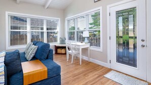 A cozy living room with a blue sofa, a wooden coffee table, and a white desk with a lamp. There are large windows with blinds, a white door with a glass panel, and a wooden ceiling.
