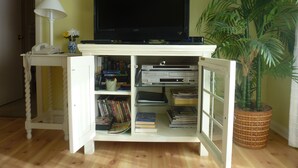 Un espacio habitable pequeño con una televisión de pantalla plana sobre un mueble blanco, un escritorio con lámpara y una planta en maceta.