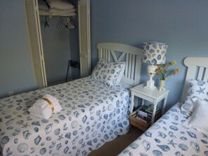 Un dormitorio con dos camas, cada una con cubrecama estampado con conchas azules y blancas. Entre las camas hay una mesita de noche blanca con una lámpara y un florero.