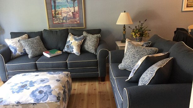 Una sala de estar con un sofá gris oscuro, un taburete estampado y cojines con motivos florales. Un cuadro enmarcado de una playa cuelga en la pared, y una lámpara está sobre una mesita.