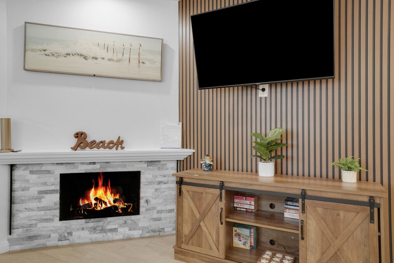 Un salon avec une cheminée, un téléviseur fixé au mur, un meuble en bois et une enseigne «Beach» au-dessus du manteau de cheminée.