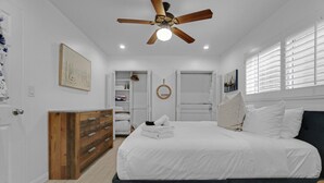 Une chambre à coucher comprenant un lit, une commode en bois, un ventilateur de plafond, un placard et une fenêtre avec des stores.