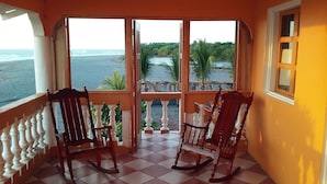 Un balcón con dos mecedoras, piso de baldosas y vista a la playa y palmeras.