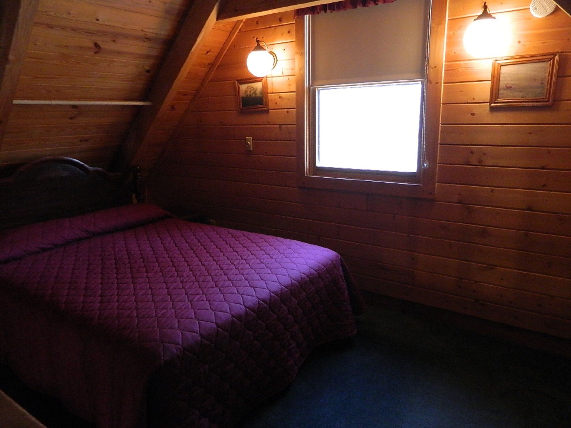 Een kamer met een houten schuine plafond, een bed met een paars sprei, een raam met een witte jaloezie en twee wandlampen.