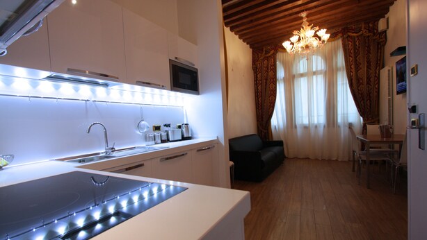 A modern kitchen with a built-in stove, microwave, and sink. There is a dining area with a table and chairs, and a living room with a sofa. A chandelier hangs from the ceiling. - SPLENDID STAR SAN MARCO SQUARE - Magic Dream ! All Inclusive Options-WIFi (Venice)