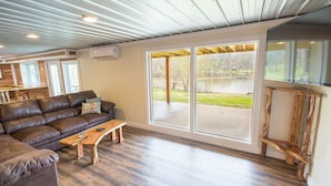 A spacious living room with a leather sofa, wooden coffee table, and large windows offering a view of a backyard. - Spacious Lake Barkley Retreat, 7 Queen Beds, Peaceful Cul-de-Sac , Dock Access (Cadiz)
