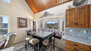 A kitchen with a wooden ceiling, a dining table with bar stools, a large window, and a view of the outdoors. - Lake Arrowhead Luxury Escape - Amazing Views For Families, Pets & Remote Workers (Lake Arrowhead)