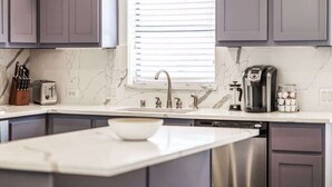 A modern kitchen with grey cabinets, a white countertop, a built-in dishwasher, a coffee maker, and a window with blinds. - Driem House Chic + Legacy Shops Walkable (Plano)