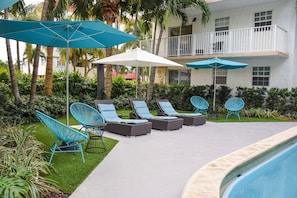 A poolside area with lounge chairs and umbrellas, surrounded by greenery and a building with balconies.