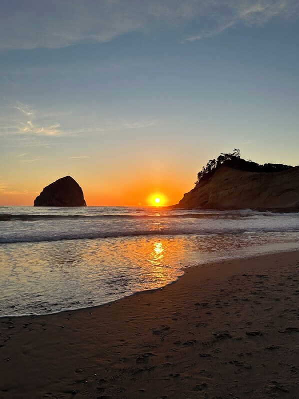A beach with a large rock formation and a cliff with vegetation, reflecting the sunset.